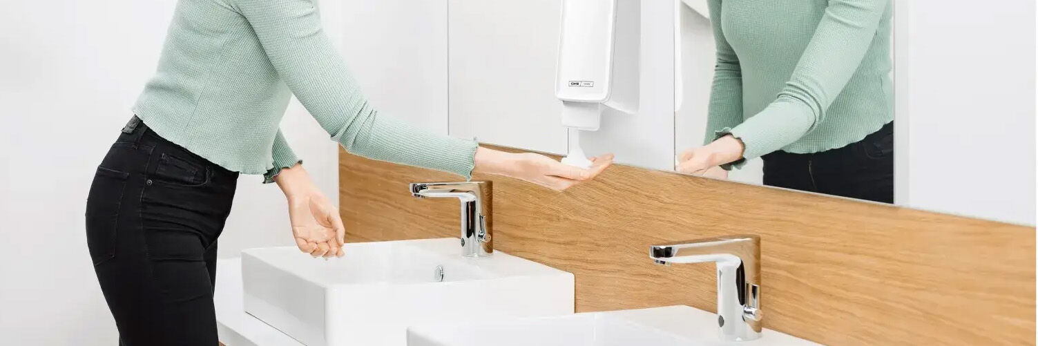 frau h&auml;lt hand unter seifenspender in &ouml;ffentlichem wc mit zwei lavabos