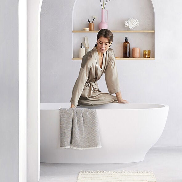 frau in seidenbademantel sitzt neben badewanne in weissem modernem bad